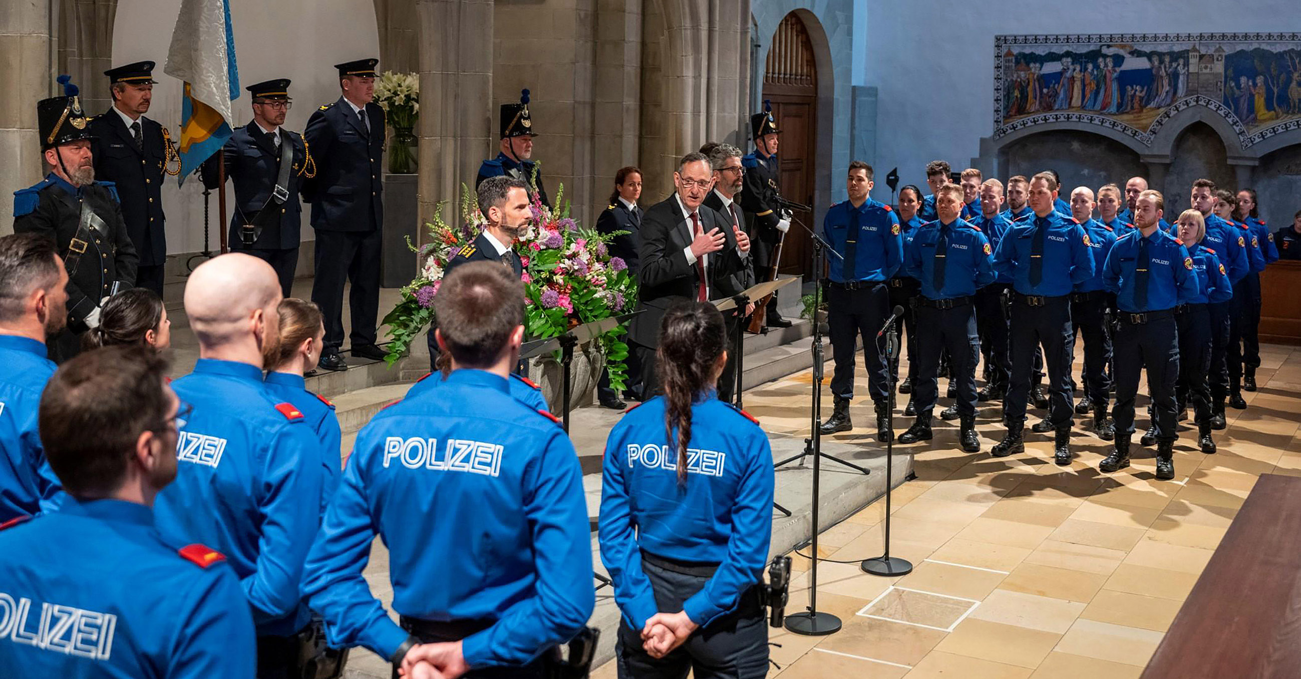 Mario Fehr hält eine Ansprache bei der Vereidigung von Polizeiaspirantinnen und Polizeiaspiranten im Fraumünster. Neben ihm stehen Kantonspolizei-Kommandant Marius Weyermann und, in Polizeiuniform, die Aspirantinnen und Aspiranten. 