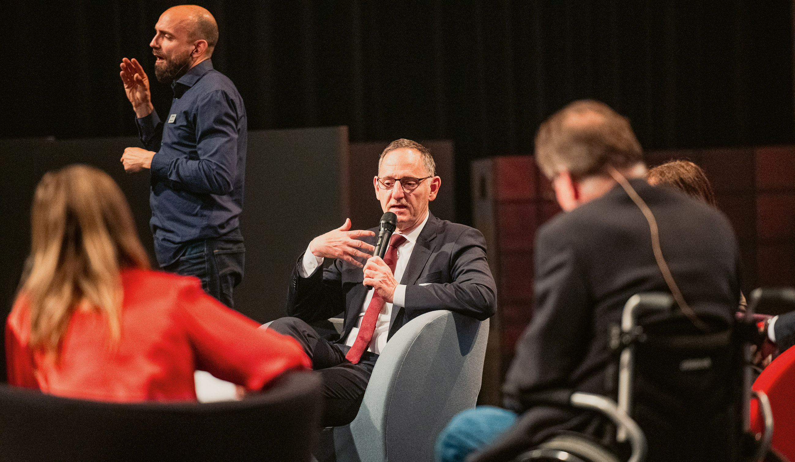 Mario Fehr sitzt auf dem Podium des Eröffnungsanlasses der Aktionstage Behindertenrechte 2024 in Zürich und spricht. Im Hintergrund verdolmetscht ein Mann das Gesagte in Gebärdensprache. Daneben sieht man auf dem Podium von hinten einen Mann, der im Rollstuhl sitzt und eine Frau mit einem roten Gilet.