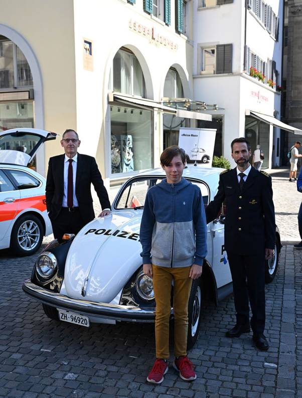 Mario Fehr, Kapo-Kommandant Marius Weyermann und der Schützenkönig des Knabenschiessens 2023, Miro Scheiwiller, stehen neben einem klassischen Polizeiauto und lachen in die Kamera. 