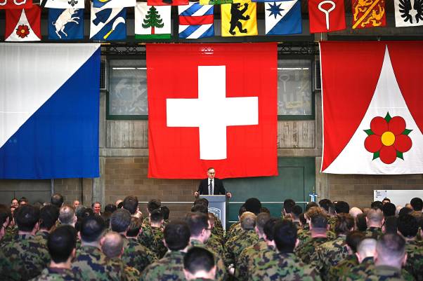 Mario Fehr befindet in einer Halle am Rednerpult, vor ihm stehen dutzende Personen in Militär-Uniform. Hinter ihm hängen verschiedene Fahnen, darunter eine riesige Schweizer Flagge.