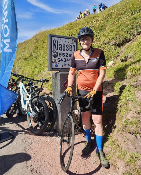Mario Fehr steht in kurzen Hosen und Shirt mit seinem Bike vor der «Klausen»-Ortstafel. Im Hintergrund befindet sich ein grüner Hügel mit weiteren Personen. 
