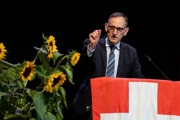 Mario Fehr steht im schwarzen Anzug am Rednerpult, schaut in das Publikum und spricht. Vor ihm hängt eine Schweizerflagge, links von ihm befinden sich mehrere Sonnenblumen. 