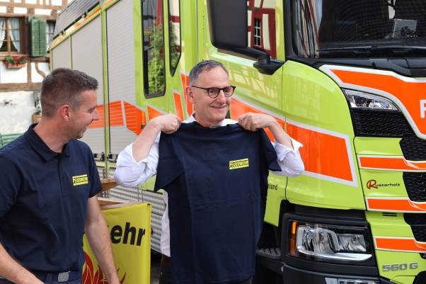 Mario Fehr hält ein schwarzes Feuerwehr-T-Shirt in die Kameras und lacht. Links von ihm steht Kommandant Peter Stutz. Hinter den beiden befindet sich das Grosslöschfahrzeug.