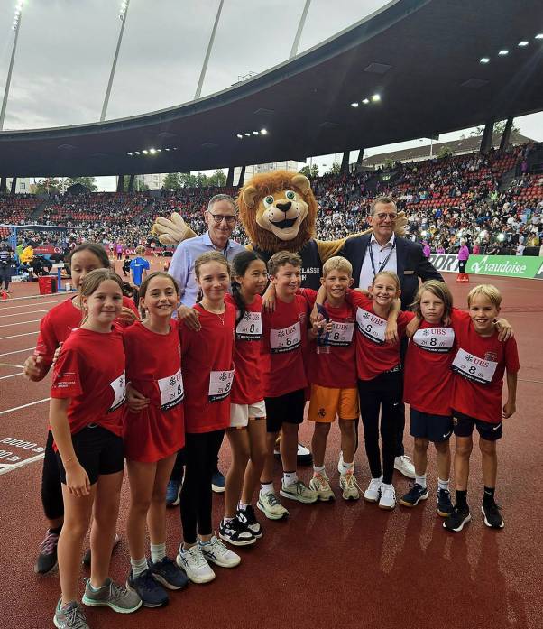 Mario Fehr steht zusammen mit dem Maskottchen, dem Löwen Leo Letzi, und den Brütten Allstars im Stadion Letzigrund. Die Mädchen und Jungs tragen rote T-Shirts und weisse Startnummern. Alle lachen in die Kamera. Im Hintergrund ist die gut gefüllte Zuschauertribüne zu sehen. 