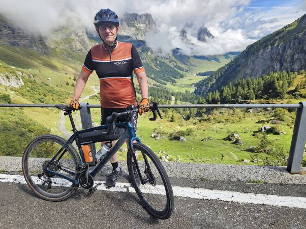 Mario Fehr steht mit seinem Bike am Strassenrand. Er trägt ein oranges Shirt, schwarze Radlerhosen, orange Handschuhe und einen schwarzen Velohelm. Im Hintergrund ist die Aussicht auf grüne Wiesen, Bäume und Berge zu sehen.