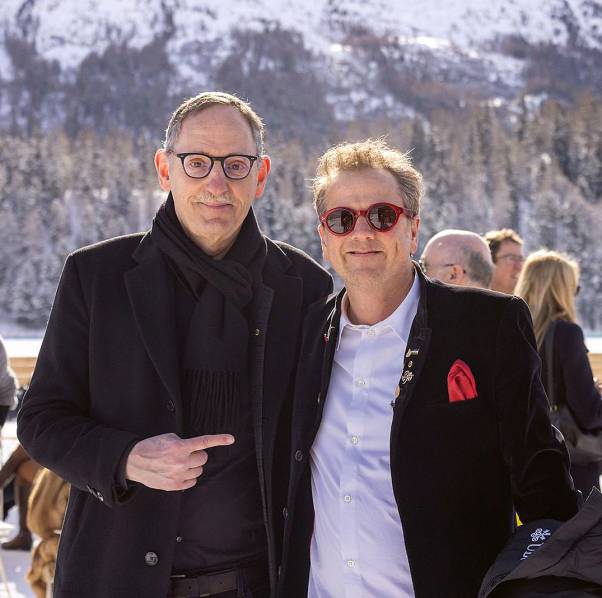 Mario Fehr ist mit einer schwarzen Jacke gekleidet. Er steht neben Christian Jott Jenny, der ein blaues Hemd und einen schwarzen Anzug trägt. Hinter ihnen ist ein Berg mit Wald und Schnee zu sehen. 