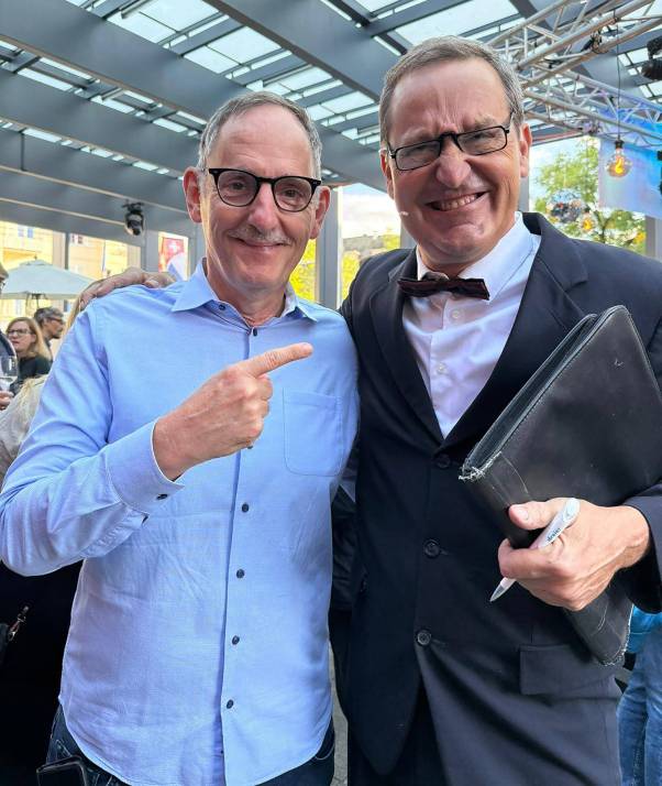 Mario Fehr steht neben Peter Winkler. Fehr lacht und trägt ein hellblaues Hemd. Winkler spielt seine Figur Theo Hitzig. Er ist bekleidet mit einem schwarzen Jackett und einem schwarzen Schlips. In der Hand hält er eine Mappe und einen Stift und er schaut freudig-zerknirscht. 