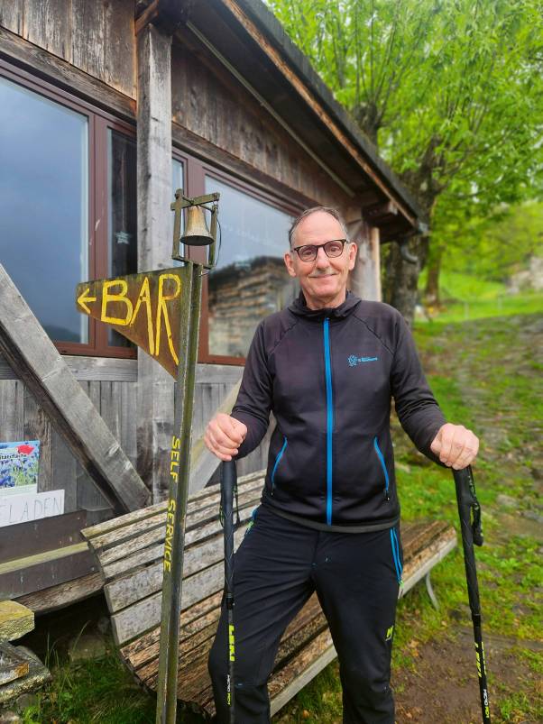 Mario Fehr steht vor einem Holzhaus. Er hält zwei Wanderstöcke in der Hand und trägt eine Trainerjacke des Sportamts des Kantons Zürich. Im Hintergrund sind Bäume zu sehen. Neben ihm steht ein Wegweiser mit der Aufschrift «Bar» und einem Pfeil nach links. 