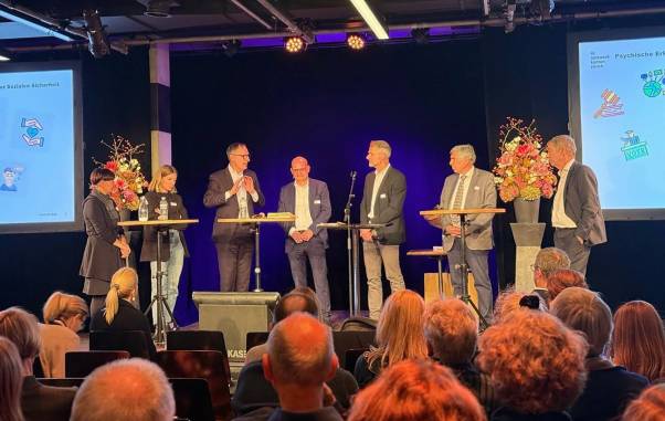 Mario Fehr steht auf einer Bühne und ist im Gespräch mit verschiedenen weiteren Personen. Vor der Bühne sind diverse Menschen von hinten zu sehen, die zuhören. 