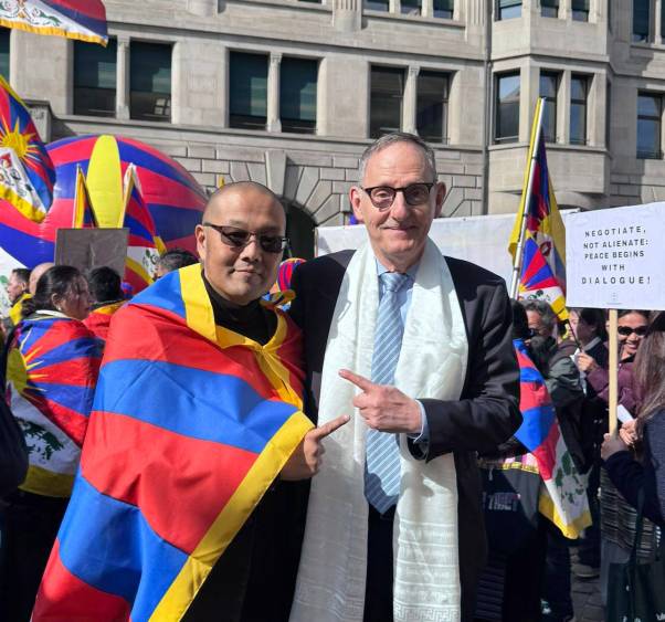 Mario Fehr steht an der Kundgebung neben Namgyal Gangshontsang. Sie zeigen mit den Fingern gegenseitig auf einander und lächeln. Hinter ihnen sind tibetische Fahnen zu sehen, die Menschen in die Luft halten. 